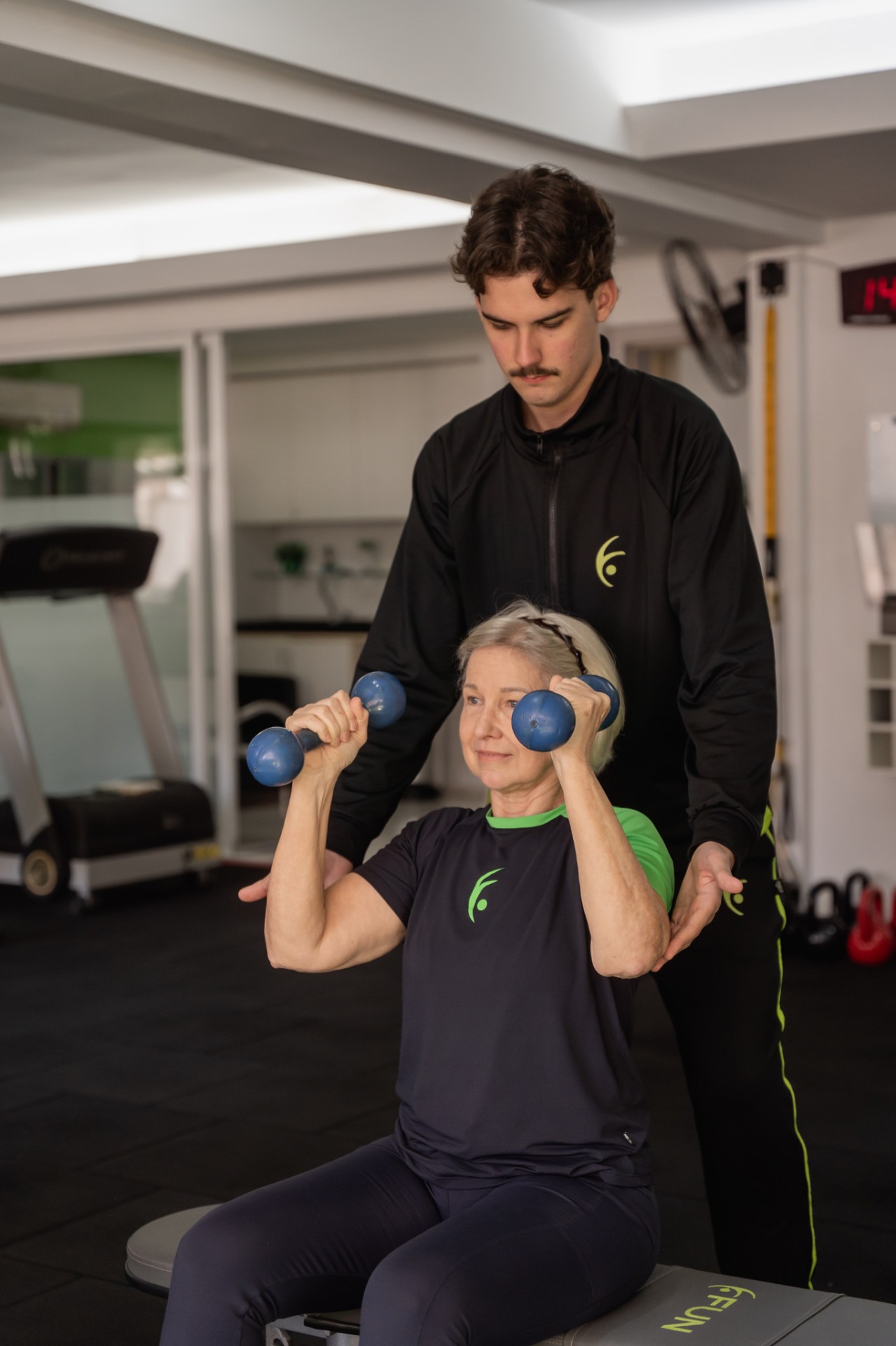Aluna fazendo exercício com halteres, acompanhada pelo professor da Fun